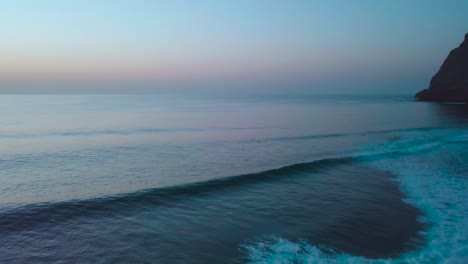 Aerial-drone-shot-of-small-breaking-waves-along-mountainous-coastline-with-sunset-afterglow