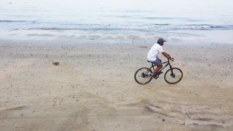 A-young-Brown-Indian-Man-cycling-early-morning-on-beach-shore-drone-shot-aerial-view-video-background,-A-young-Indian-man-doing-exercise-on-beach-shore