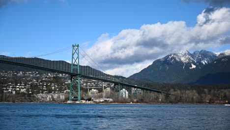 Zeitraffer-Von-Kleinen-Yachten,-Die-In-Richtung-Vancouver-Segeln,-Während-Ein-Schlepper-Zum-Meer-Fährt-Und-Autos-über-Die-Lions-Gate-Bridge-Fahren,-Während-Wolken-über-Den-Schneebedeckten-Felsigen-Berg-Ziehen
