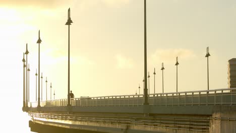 Golden-Hour-Sunlight,-Bridge-Traffic-and-Silhouette-of-a-Person-Riding-Electric-Scooter,-Static-Shot