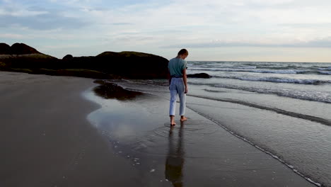 Young-girl-walking-toward-ocean-waves-on-sandy-coastline,-back-view