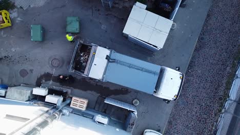 Luftaufnahme-Eines-Müllwagens,-Der-Müll-Automatisch-Belädt,-Menschen,-Die-Müllcontainer-Verschieben,-Weitwinkel-Drohnenaufnahme-Aus-Der-Vogelperspektive,-Die-Sich-Langsam-Nach-Links-Dreht