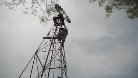 Man-climbing-a-windmill-in-a-farm-in-northern-Mexico,-low-angle-wide-shot