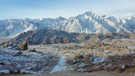 Majestic-gravel-road-leading-to-snowy-mountain-range-during-snowfall,-aerial-fly-forward-view