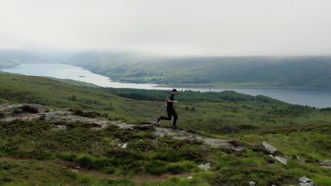 Aerial-Tracking-View-Of-Male-Running-Along-Ridge-Of-Ullapool-Hill-With-Loch-Achall-In-The-Background