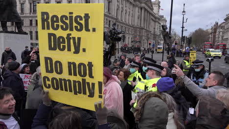 Police-officers-are-surrounded-as-they-try-and-talk-to-Coronavirus-conspiracy-anti-vaccination-protestors,-one-of-whom-holds-cup-a-yellow-placed-that-says-Resist,-defy,-do-not-comply