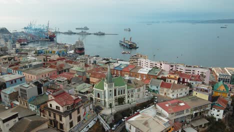 Aerial-dolly-in-of-colorful-houses-and-Lutheran-Church-leading-to-Valparaiso-Sea-Port-and-ships-docked,-Chile