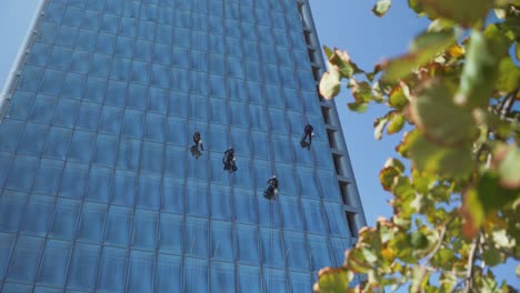 Close-up-of-four-window-cleaners-hanging-and-swinging-from-a-glass-curtain-wall-of-modern-skyscraper-at-daytime,-Santiago,-Chile