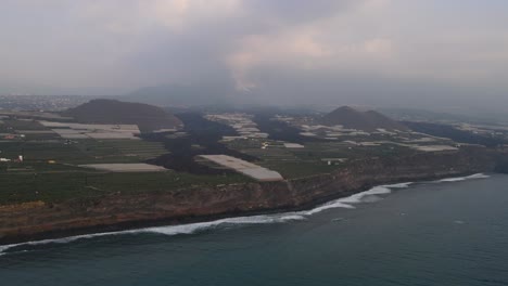 Aerial-towards-solidified-lava-on-banana-plantation-and-volcano-shrouded-in-thick-smoke-in-background,-La-Palma-island