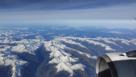 Looking-through-the-window-aircraft-during-flight-and-seeing-a-snow-covered-European-Alps-mountain-range-with-blue-sky-without-clouds