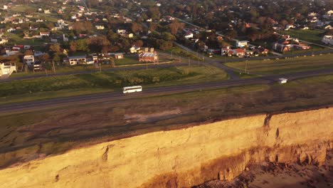 Aerial-tracking-shot-of-white-bus-driving-on-coastal-road-beside-cliffs-and-ocean-during-sunset