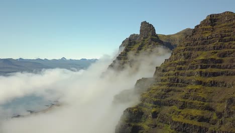 Luftaufnahme-über-Einem-Grünen,-Steilen-Hügelgipfel,-Wolken-Bedecken-Die-Berge-Im-Hintergrund,-Tagsüber-In-Der-Kleinen-Stadt-Djúpivogur-In-Der-Nähe-Der-Isländischen-Fjorde