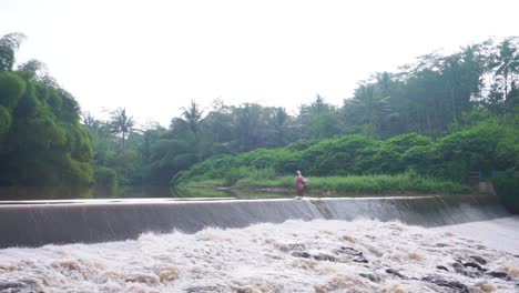 An-old-woman-crosses-a-dam-in-Magelang,-Indonesia