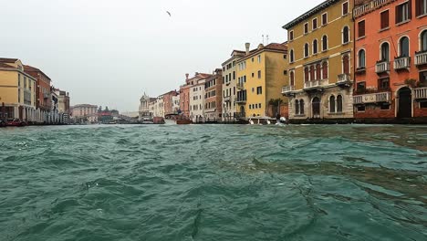 Low-angle-water-surface-pov-of-Venice-and-motorboat-passing-seen-from-navigating-boat,-Venice-in-Italy
