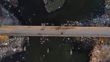 Aerial-top-down,-scooters-driving-on-bridge-over-river-polluted-with-plastic-trash
