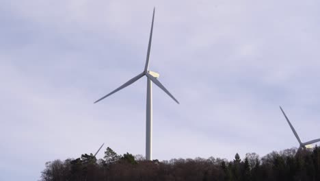One-single-windmill-in-center-of-frame-with-two-more-windmills-hidden-below-treetops-of-forest---Static-clip-of-rotating-turbines-in-Norway