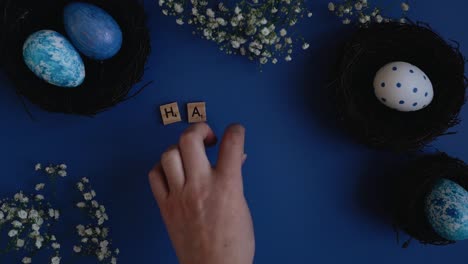 Top-view-of-hands-writing-happy-easter-with-the-help-of-scrabble-letters-on-a-blue-background-surrounded-by-beautifully-decorated-easter-eggs