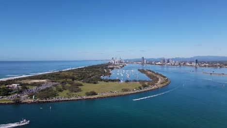High-fast-moving-drone-view-travelling-over-a-man-made-ocean-inlet-towards-an-urban-boat-harbor-with-a-metropolitan-skyline-and-mountain-range-in-the-distance