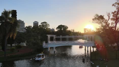 Aerial-dolly-in-of-boats-sailing-under-white-bridge-in-Rosedal-gardens-in-Palermo-neighborhood-at-sunset,-Buenos-Aires