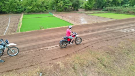 Motorcycle-Riders-Drive-On-Dirt-Road-Through-Lush-Fields-In-Cambodia