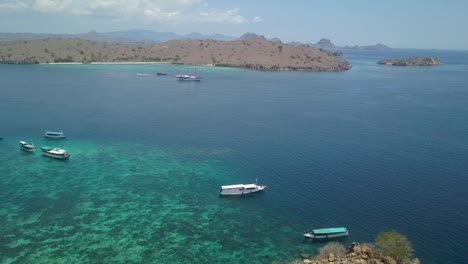 Tourist-boats-moored-on-Idyllic-bay-encircled-by-Pink-Beach-Komodo-Island,-Indonesia---Wide-Aerial-Panoramic-shot