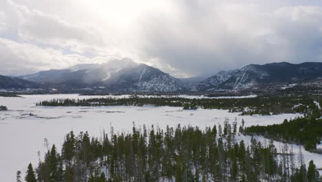 Aerial-moving-left-and-up-to-reveal-icy,-frozen-lake-surrounded-by-snow-and-green-pine-trees-with-large-snowy-ski-town-mountains-in-the-background-near-Silverthorne-and-Frisco-Colorado