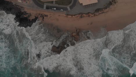 Aerial-top-down-pov-of-rough-ocean-and-waves-crashing-on-beach,-Ponta-da-Calheta