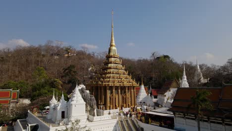Wat-Phra-Phutthabat,-Saraburi,-Thailand,-an-eye-level-aerial-shot-towards-the-stairs-going-up-the-temple-and-brown-dying-trees-at-the-background,-people-have-come-to-pray-and-seek-blessings