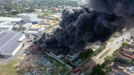Black-smoke-cloud-coming-out-of-warehouse-fire,-shot-with-drone