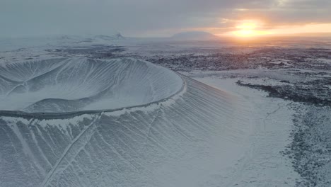 Breathtaking-drone-footage-of-snowy-tephra-cone-or-tuff-ring-volcano-during-beautiful-sunrise-in-background---Cold-frozen-winter-day-on-Iceland-showing-Hverfjall-Volcano-system-reserve