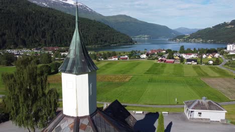 Forward-moving-aerial-passing-really-close-to-tower-of-Loen-church---Showing-church-building-with-panoramic-view-over-green-fields-and-Loenfjord-in-background-during-summer---Norway