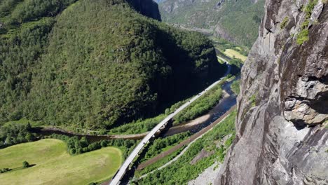 Abwärtsbewegende-Antenne-Entlang-Der-Bergwand-–-Blick-Auf-Die-Norwegische-Autobahn-E-16-Zwischen-Bergen-Und-Voss-Mit-Eisenbahn-Und-Lachsfluss-An-Den-Seiten-–-Spektakuläre-Landschaft