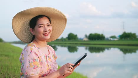 Porträt-Einer-Schönen-Asiatischen-Frau-Mit-Hut,-Die-Mit-Einem-Tragbaren-Gerät-In-Die-Kamera-Blickt