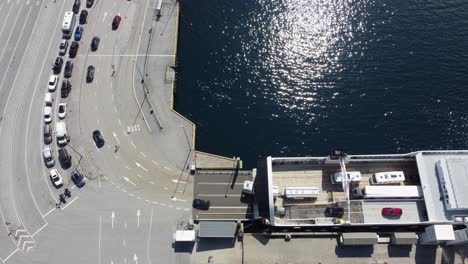 Norwegian-huge-ferry-Mf-Oppedal-discharging-cars-in-Lavik-ferry-harbour-at-sunny-summer-day---Line-of-cars-waiting-to-get-onboard---Static-top-down-aerial-showing-ferryboat-and-road-beside-sea