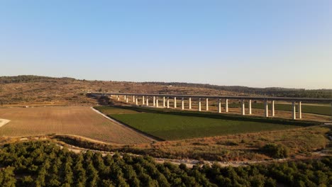 Vista-Aérea-De-Un-Largo-Puente-De-Carretera-Sobre-Un-Paisaje-Con-Campos-Y-Prados-Al-Atardecer-En-Israel