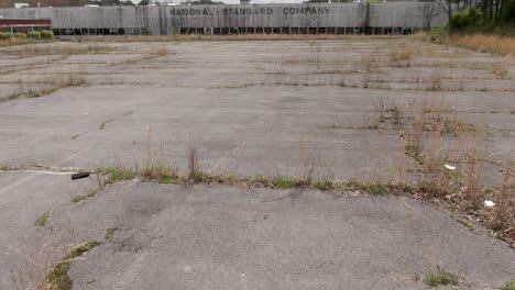 Drone-Footage-Panning-Up-and-Sliding-Over-an-Overgrown-Parking-Lot-Towards-an-Abandoned-National-Standard-Factory