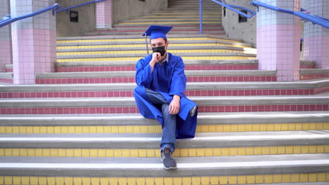 Male-graduate-sits-at-school-in-his-cap-and-gown-with-face-mask-on