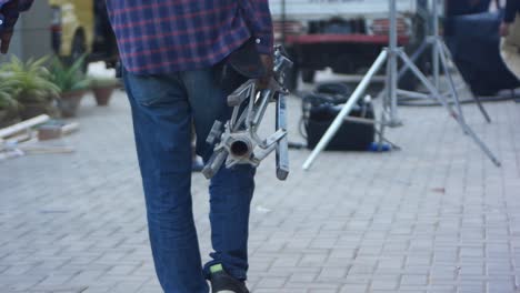 Male-Crew-Carrying-Metal-Tripod-Stand-On-Set