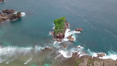 Drone-shot-of-Coral-island-overgrown-by-green-trees-that-are-buffeted-by-the-waves-crashing-in-the-tropical-ocean