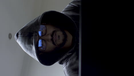 A-vertical-video-of-a-hacker-working-on-his-laptop-computer-wearing-a-dark-hoodie-covering-his-head,-the-man-planning-his-next-cyber-attack-as-he-works-late-into-the-night