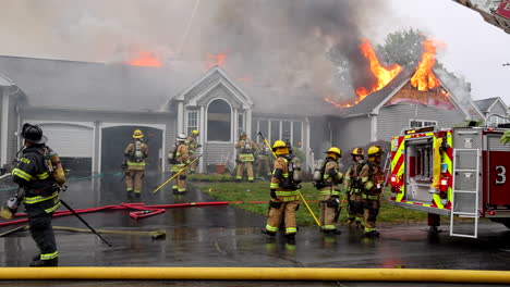Feuerwehrleute-Schlagen-Das-Fenster-Eines-Hauses-Ein,-Das-In-Flammen-Steht-Und-Dunkle-Rauchwolken-Vom-Dach-Aufsteigen