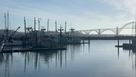 Fishing-Boats-Anchored-At-The-Port-At-Yaquina-Bay-With-Arch-Bridge-In-The-Distance-In-Newport,-Oregon