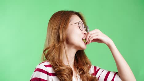 Fröhliche-Junge-Asiatische-Frau-In-Gestreiftem-T-Shirt-Mit-Brille-Genießt-Es,-Frische-Rote-Erdbeeren-Zu-Probieren-Und-Zu-Essen,-Grüner-Hintergrund