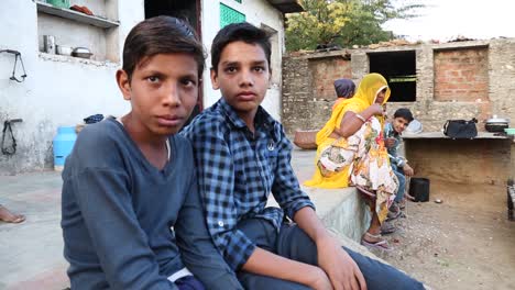 Zwei-Indische-Dalit-Kinder-Aus-Dem-Ländlichen-Dorf-Rajasthan-In-Indien-Schauen-In-Die-Kamera,-Im-Hintergrund-Ihre-Familie
