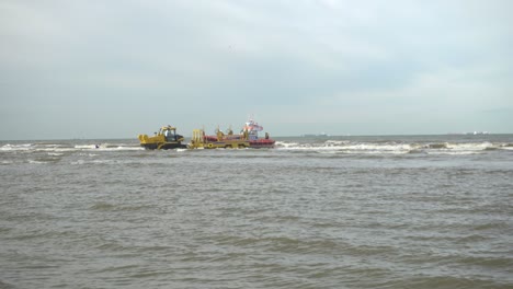 Mann-Im-Boot-Paddelt-In-Richtung-KnRM-Rettungsboot,-Das-An-Einem-Traktor-Im-Meer-Mit-Tosenden-Wellen-Bei-Flut-In-Den-Niederlanden-Befestigt-Ist