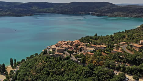 Sainte-Croix-du-Verdon-France-Aerial-v4-establishing-shot,-drone-fly-around-hillside-village-overlooking-beautiful-lake-of-sainte-croix-with-vivid-turquoise-blue-water-surrounded-by-hills---July-2021