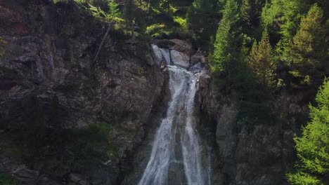 Alpenwasserfall-An-Einer-Felswand-Mit-Kiefernwald,-Luftaufnahme-Bei-Sonnenuntergang