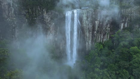 Cinematic-view-looking-through-rising-mist-revealing-a-majestic-waterfall-spilling-over-a-lush-rainforest-covered-mountain