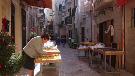 Anciana-En-Un-Puesto-Callejero-Vendiendo-Productos-En-Una-Calle-Estrecha-Y-Hermosa-En-Bari,-Italia