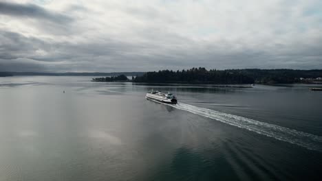 Following-behind-a-large-commuter-ferry-on-dark-calm-water,-gloomy-clouds-reflect,-aerial